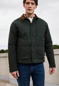 Dunkelgrüne Jacke mit einem braunen Cordkragen, ausgestattet mit einem Knopfverschluss und zwei großen Vordertaschen, getragen mit blauen Jeans.
