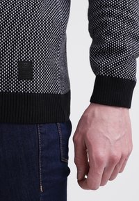 Stickad svart och vit tröja med ett strukturerat mönster, ribbade svarta ärmslut och en svart etikett på nedre vänstra sidan. Bärs med mörka jeans.