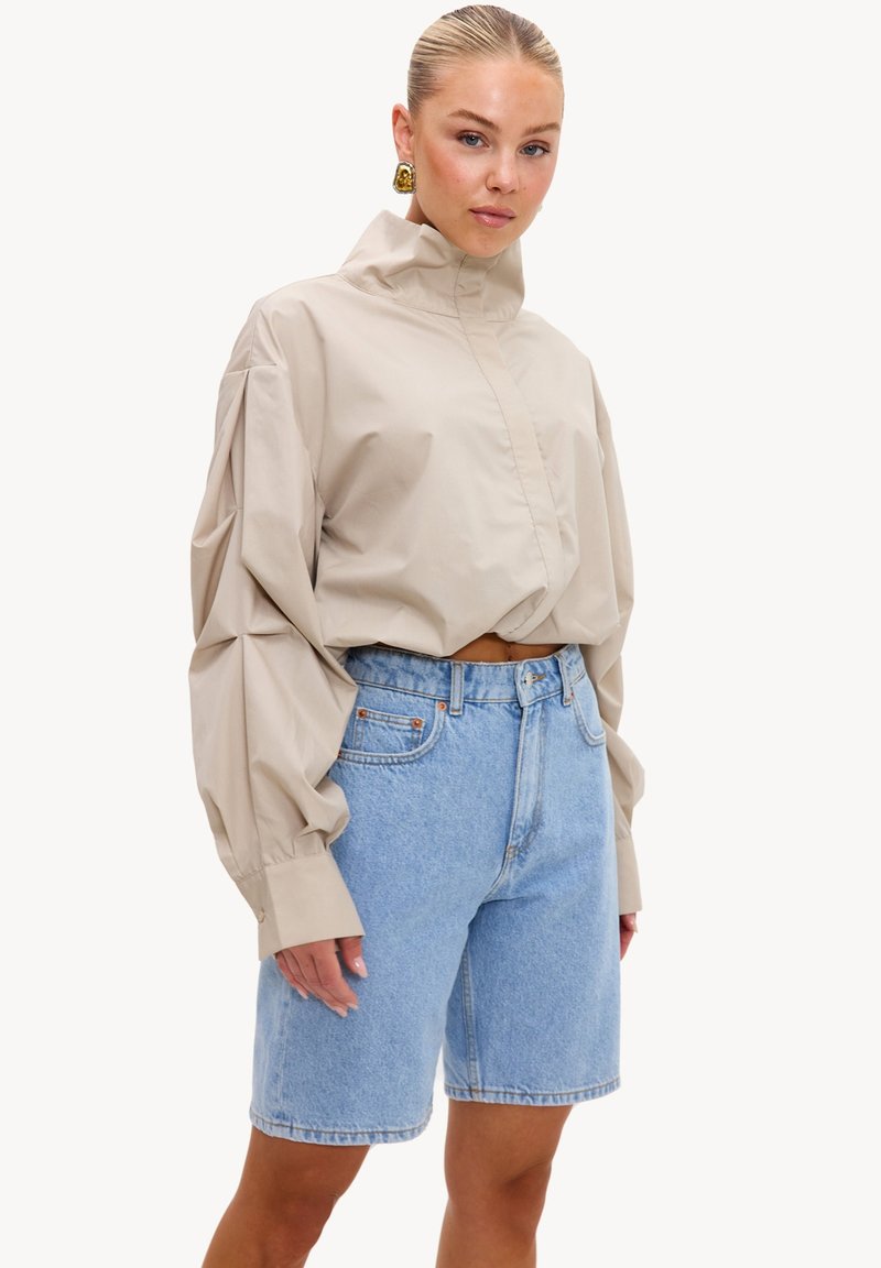 Femme aux cheveux plaqués en arrière portant un chemisier beige oversize à col haut et un short en jean bleu clair longueur genoux.