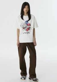 Witte t-shirt met korte mouwen met een kleurrafische afbeelding van een hart, een roos en tekst. Gecombineerd met losse, bruine, gestructureerde broek en gestreepte schoenen.