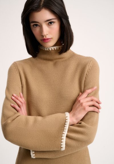 Pull en tricot beige avec un col haut, présentant des accents froncés blancs au col et aux poignets ; coupe décontractée avec des manches longues.