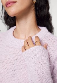 Pull tricoté violet clair avec une texture en maille lâche et des poignets côtelés. Des bagues dorées portées aux doigts ajoutent un détail subtil à la tenue.