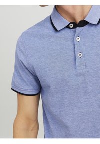 Detailní záběr na osobu, která má na sobě světle modrou polo košili s texturou, černým lemem na límci a rukávech a třemi bílými knoflíky na légě.