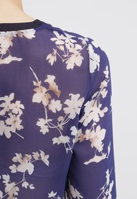 Blouse transparente avec un motif floral beigne et blanc sur fond bleu foncé. Tissu doux avec un col rond et des manches longues.