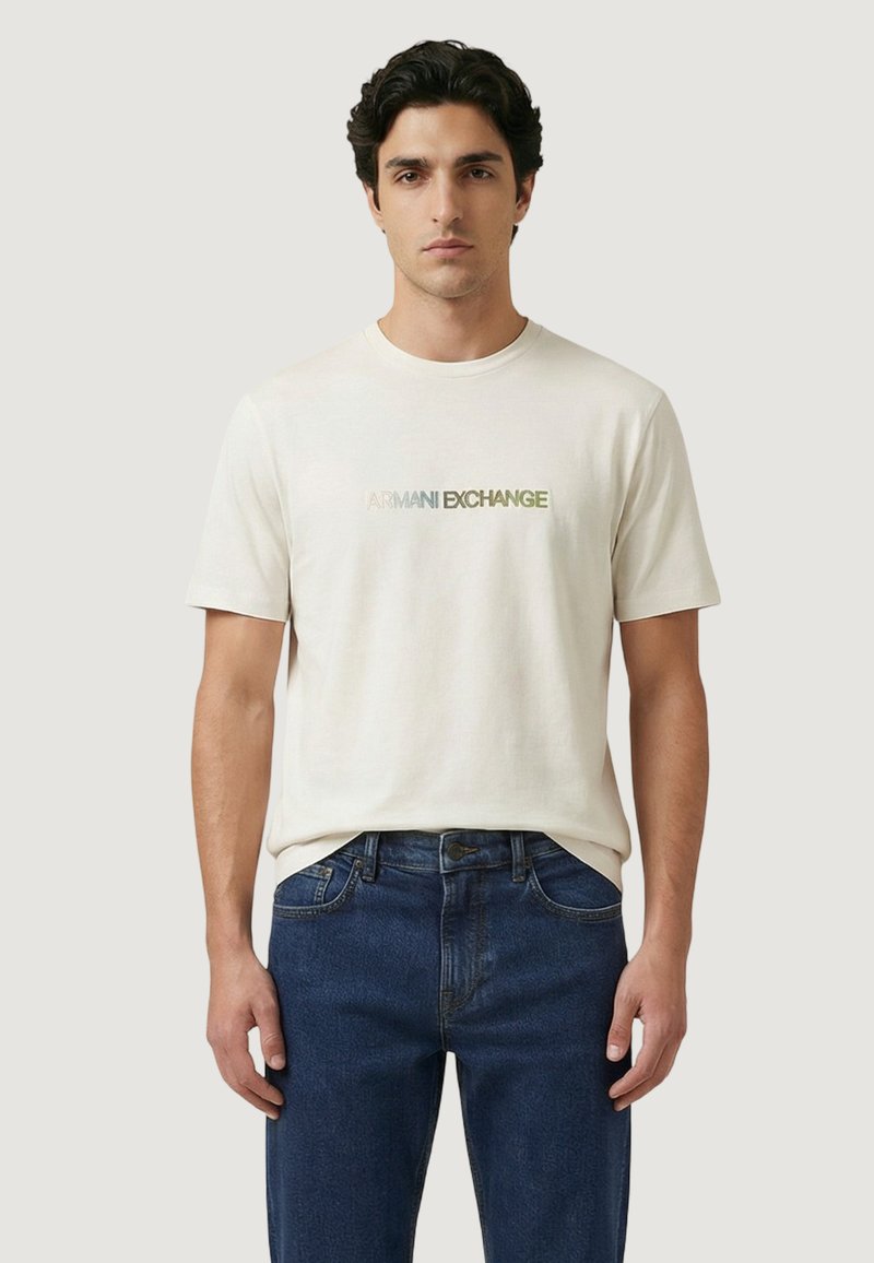Homme portant un t-shirt blanc Armani Exchange et un jean bleu, debout devant un fond clair uni.