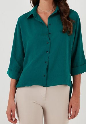 Femme portant une chemise boutonnée bleu sarcelle aux manches retroussées et un pantalon beige, avec de longs cheveux bruns et un fin collier en or visible.