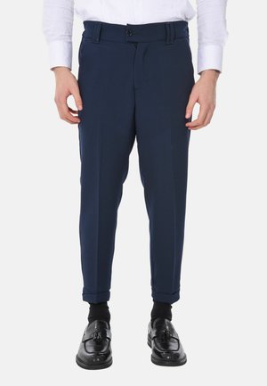 Pantaloni sartoriali blu navy con una texture liscia, dotati di chiusura a bottone, passanti per cintura e orli arrotolati, abbinati a mocassini neri e calzini.
