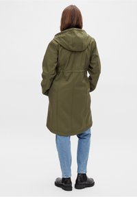 Manteau d'extérieur vert olive avec capuche, taille resserrée et coupe droite, associé à un jean bleu clair et des bottines noires.