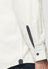 Witte lange mouwen shirt met gestructureerde stof, donkerblauwe accenten op de manchetten en zijslitsen, en een knoopsluiting op de manchetten.