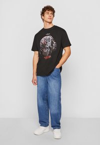 Schwarzes Baumwoll-T-Shirt mit einem grafischen Design eines Kopfes in roten Akzenten, kombiniert mit hellblauen geraden Jeans und weißen Sneakern.
