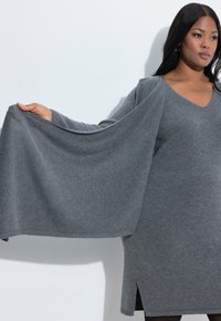 Vestido de punto gris con escote en V, mangas largas y aberturas laterales. La tela tiene una textura suave y cae con gracia.