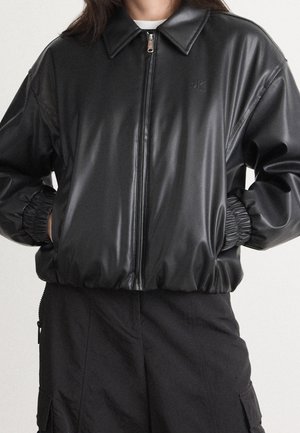 Person trägt eine schwarze Lederjacke mit Reißverschluss, elastischen Bündchen und Kragen, Hände in den Taschen, kombiniert mit schwarzen Cargohosen.