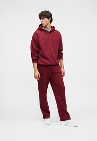 Fiatal férfi bordó kapucnis pulóverben és hozzáillő melegítőnadrágban, fehér tornacipővel, egy sima háttér előtt állva, oldalra nézve.