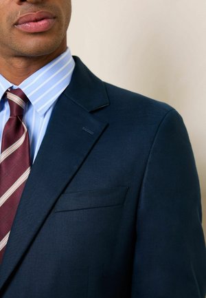 Mann trägt blaues Nadelstreifenhemd, bordeaux gestreifte Krawatte und dunkelblaue Anzugjacke vor einfarbigem Hintergrund.