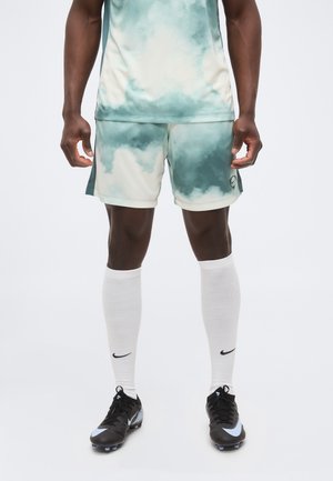 Athlète masculin portant une tenue de sport verte et blanche à motif de nuages, des chaussettes blanches montantes avec un logo Nike noir, et des crampons noirs avec des accents bleus.