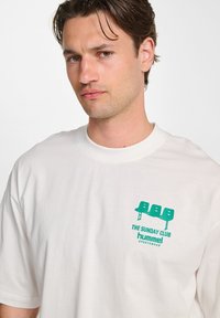 White cotton t-shirt featuring a green graphic print of three chairs and text. Crew neck, relaxed fit, with short sleeves.