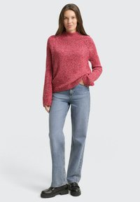 Frau steht mit der Hand in der Tasche, trägt einen bordeauxroten Strickpullover, hellblaue Jeans und schwarze Plateauschuhe vor einem hellgrauen Hintergrund.