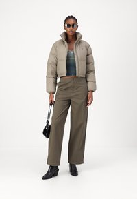 Puffer-jakke i taupe med cropped design, kombinert med olivengrønne vide bukser. Modellen har på seg en mørkegrønn topp og svarte støvler, mens hun holder en svart veske.