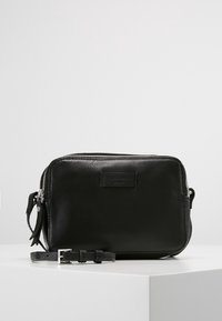 Svart läder crossbody-väska med en slät textur, dragkedjestängning, justerbar rem och en liten frametikett. Kompakt rektangulär form.