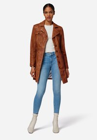 Bruine leren trenchcoat met lange mouwen, dubbelzijdige knopen en een kraag, gedragen over een wit topje en lichtblauwe skinny jeans.