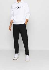 Biała bawełniana bluza z kapturem, z sznurkami i czarnym napisem logo, w połączeniu z czarnymi spodniami i białymi trampkami, charakteryzująca się prostym designem i luźnym krojem.