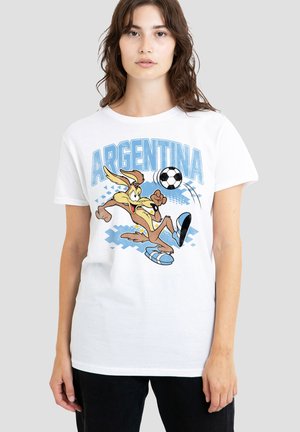 Junge Frau trägt ein weißes T-Shirt mit einem Cartoon-Kojoten, der einen Fußball tritt, und dem Wort Argentinien darüber in blauen Buchstaben.