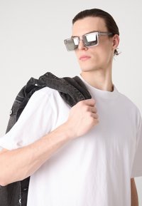 Klare rektangulære solbriller med tonede linser, båret med en vanlig hvit t-skjorte. En mørk denimjakke er slengt over skulderen.