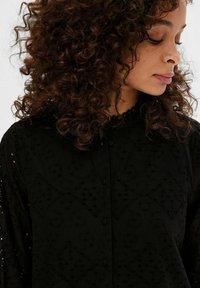 Schwarze bestickte Bluse mit Knopfleiste, gewelltem Kragen und kunstvollen Ausschnitten im gesamten Gewebe.