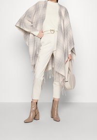 Grau- und cremefarbener kariierter Poncho mit Fransen, kombiniert mit einem cremefarbenen Pullover, hellen Hosen, beigen Stiefeletten mit Absatz und einer passenden Handtasche.