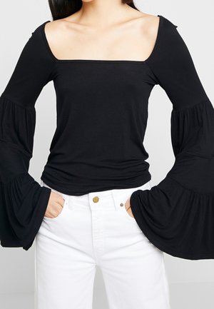 Kobieta ubrana w czarny top z odkrytymi ramionami i szerokimi dzwonkowatymi rękawami oraz białe, wysokowciskowe spodnie, z rękami w kieszeniach.