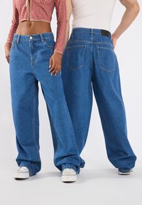 Baggy blue denim jeans with a high waist, prominent stitching, and two back pockets; worn with white canvas sneakers.