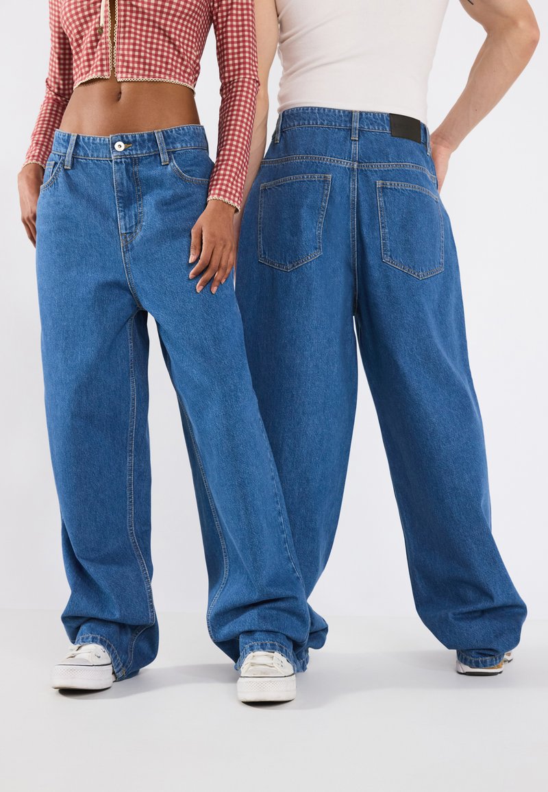 Baggy blue denim jeans with a high waist, prominent stitching, and two back pockets; worn with white canvas sneakers.