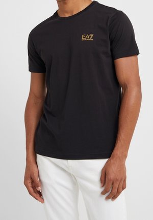 Man draagt een zwart T-shirt met korte mouwen en een gouden EA7 Emporio Armani-logo op de borst, gecombineerd met witte broek tegen een effen lichte achtergrond.
