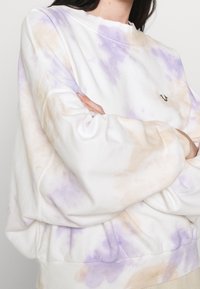 Person som bär en överdimensionerad vit sweatshirt med beige och lila batikmönster, armarna korsade, med en liten svart broderad logotyp på bröstet.