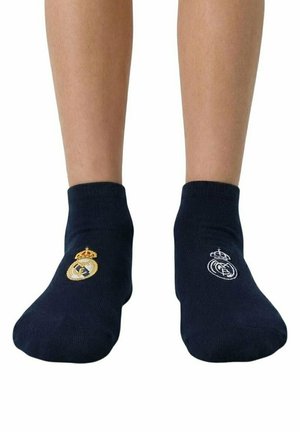 Stopy w czarnych krótkich skarpetkach, każda skarpetka ma inny haftowany złoto-biały emblemat klubu piłkarskiego Real Madrid.