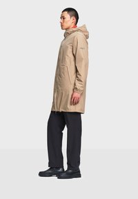 Homme portant un imperméable beige à capuche, pantalon noir et bottes noires, debout de profil sur un fond clair uni.