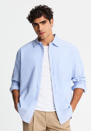 Giovane uomo con capelli ricci che indossa una camicia azzurro chiaro con bottoni sopra una t-shirt bianca e pantaloni beige, in piedi con le mani nelle tasche.