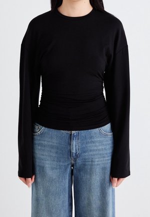 Vrouw in een strakke zwarte long-sleeve shirt met subtiele plooien aan de zijkanten en losse blauwe denim jeans, staand voor een effen witte achtergrond.