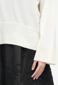 Suéter de lã creme com bainha e punhos canelados, combinado com uma saia preta com detalhes florais bordados e lantejoulas.