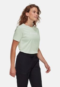 Jasnozielona koszula z krótkim rękawem i okrągłym dekoltem, z białym nadrukowanym logo z przodu, w połączeniu z czarnymi spodniami z zamkami błyskawicznymi.