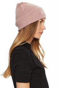 Bonnet tricoté en rose clair avec une texture côtelée; arbore un logo cousu à l'avant. Le modèle porte un haut noir à manches courtes.