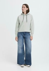 Felpa grigia chiara con coulisse nere, abbinata a jeans a gamba larga blu e sneakers nere. Design semplice, vestibilità casual e texture morbida.