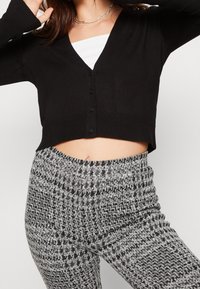 Czarny krótki kardigan z dekoltem w serek i zapięciem na guziki, zestawiony z szaro-czarnymi wzorzystymi spodniami o wysokim stanie. Gładka faktura na całej powierzchni.