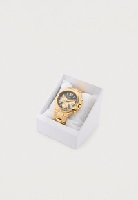 Zlata ura s črnim številčnico, ki ima tri podštevilčnice, zlate poudarke in kovinsko zapestnico. Predstavljena v beli škatli s mehko podlogo.