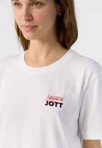 Camisa de algodón blanca con un logotipo "JOTT" multicolor bordado en el pecho. Diseñada con cuello redondo y mangas cortas.