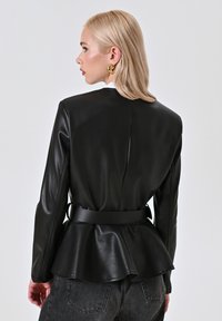 Giacca peplum nera in similpelle con maniche lunghe, vita aderente e texture liscia. Presenta uno spacco sul retro e una finitura satinata.