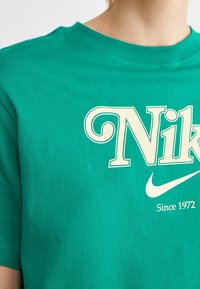 Zelené bavlněné tričko s velkým krémovým logem Nike a textem "Od roku 1972". Krátké rukávy a kulatý výstřih. Hladká textura.