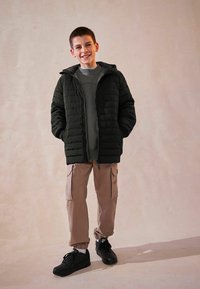 Giubbotto impermeabile nero con cappuccio, felpa grigia sotto, pantaloni cargo beige e sneakers nere. Modello in piedi contro uno sfondo chiaro.
