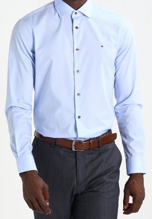 Mężczyzna ubrany w jasnoniebieską koszulę zapinaną na guziki, brązowy pasek i ciemne, w kratkę spodnie, stoi ze swobodnie opuszczonymi ramionami.