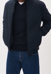 Giacca bomber navy con zip, polsini e orlo a coste. Indossata sopra un maglione scuro, abbinata a jeans in denim blu medio.
